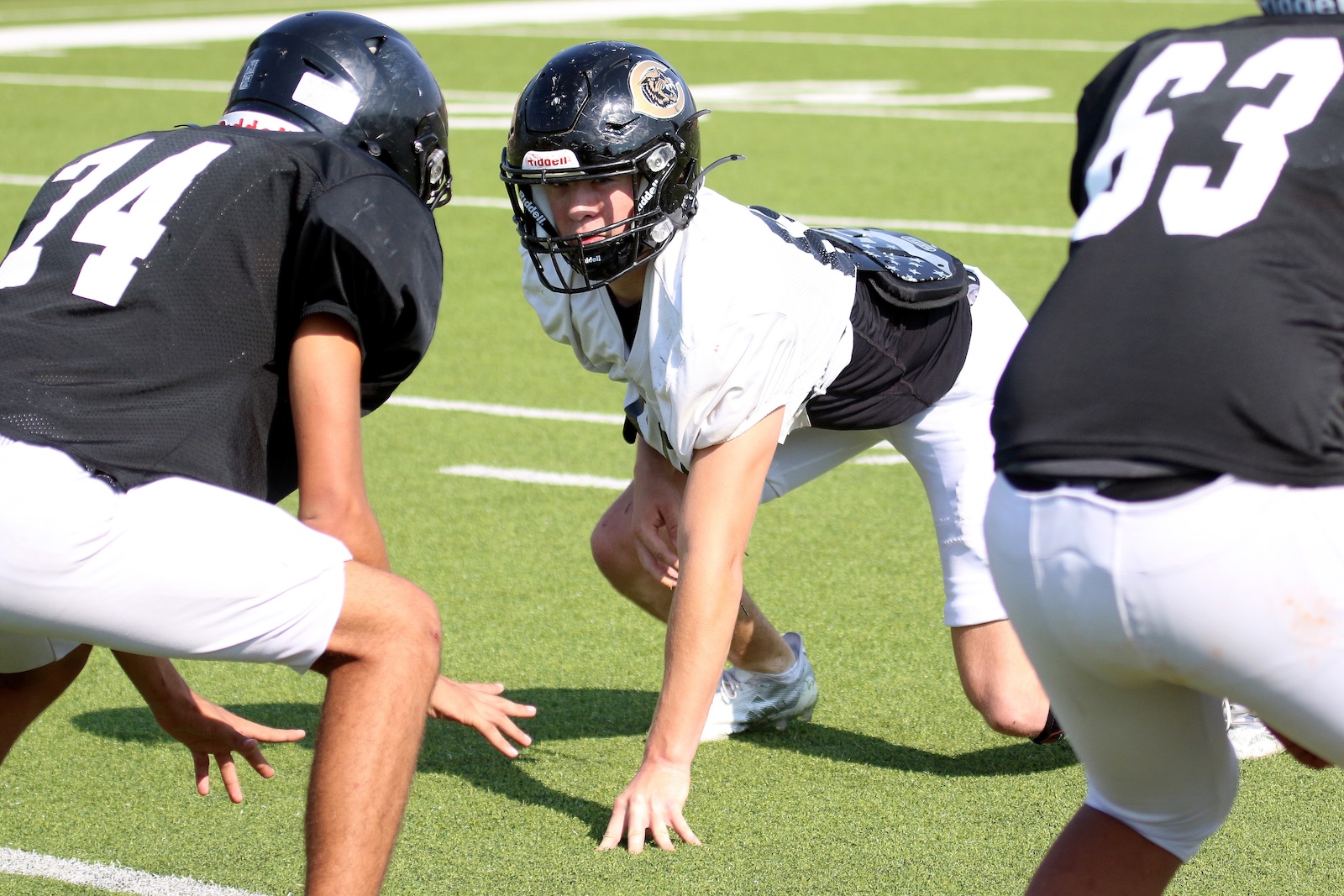 Center in a white jersey crouches at the line of scrimmage, flanked by blockers in black jerseys on a green football field at snap moment.