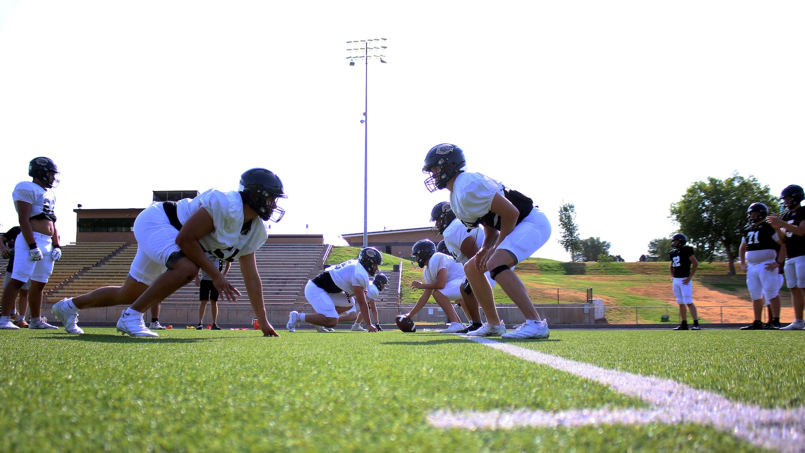 Football practice: offensive linemen crouch at the line of scrimmage facing the defense on a green field with bleachers in the background.