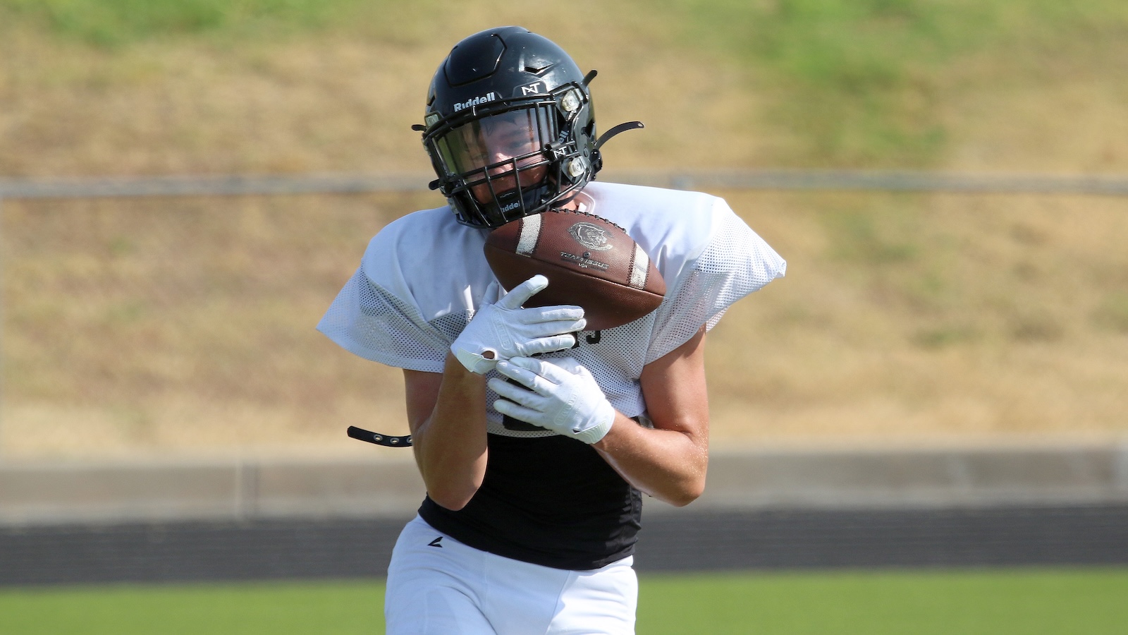 American football player in white jersey and black pants, wearing a black helmet, catching a brown football on a field.