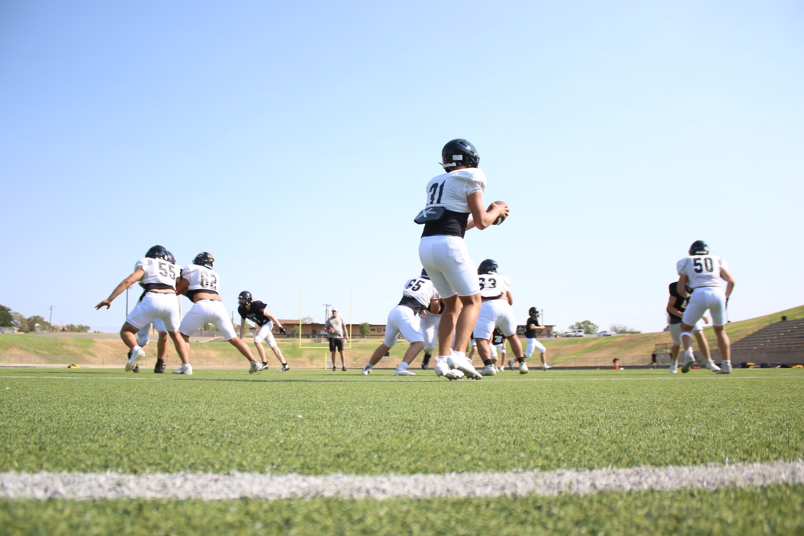 Quarterback in white jersey with ball, surrounded by teammates on a sunny football field during practice.