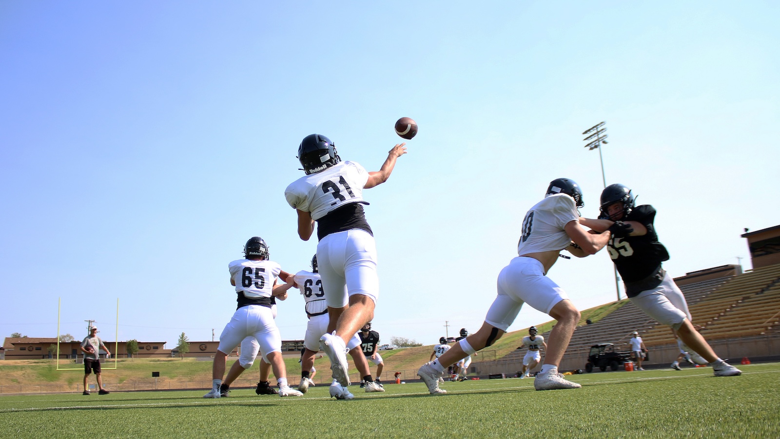 Football players in white uniforms on a sunny field; quarterback numbered 31 throws a pass while teammates block nearby.