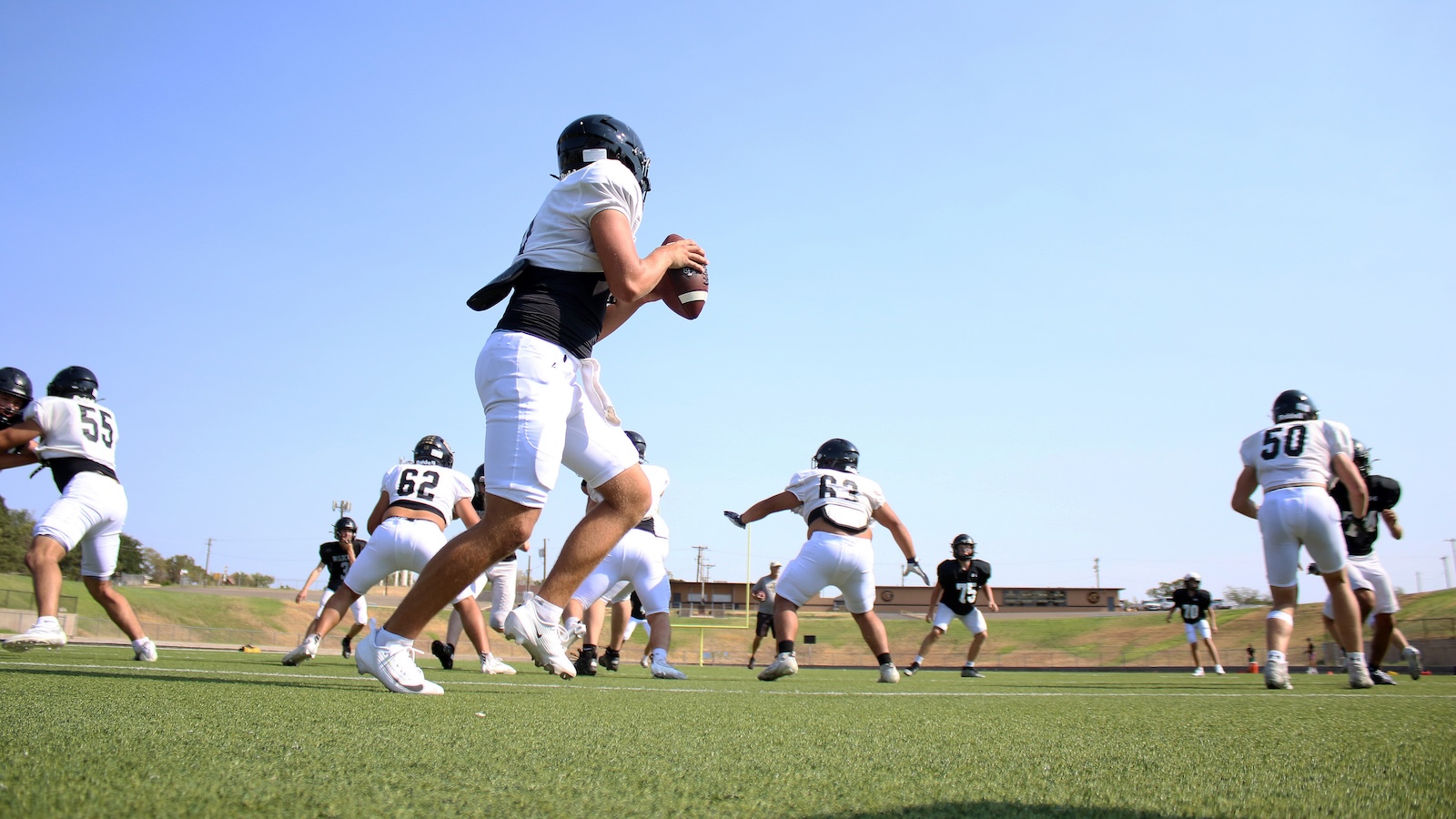 Quarterback in white uniform grips the football while teammates block during an outdoor American football play on a green field.