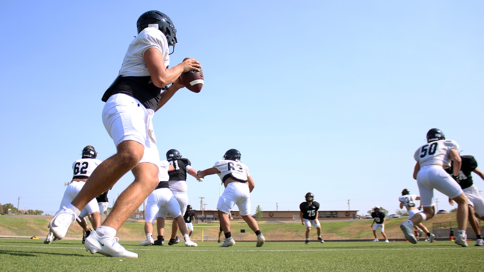 Quarterback in white jersey and helmet grips a football on a sunny practice field as teammates sprint in the background.