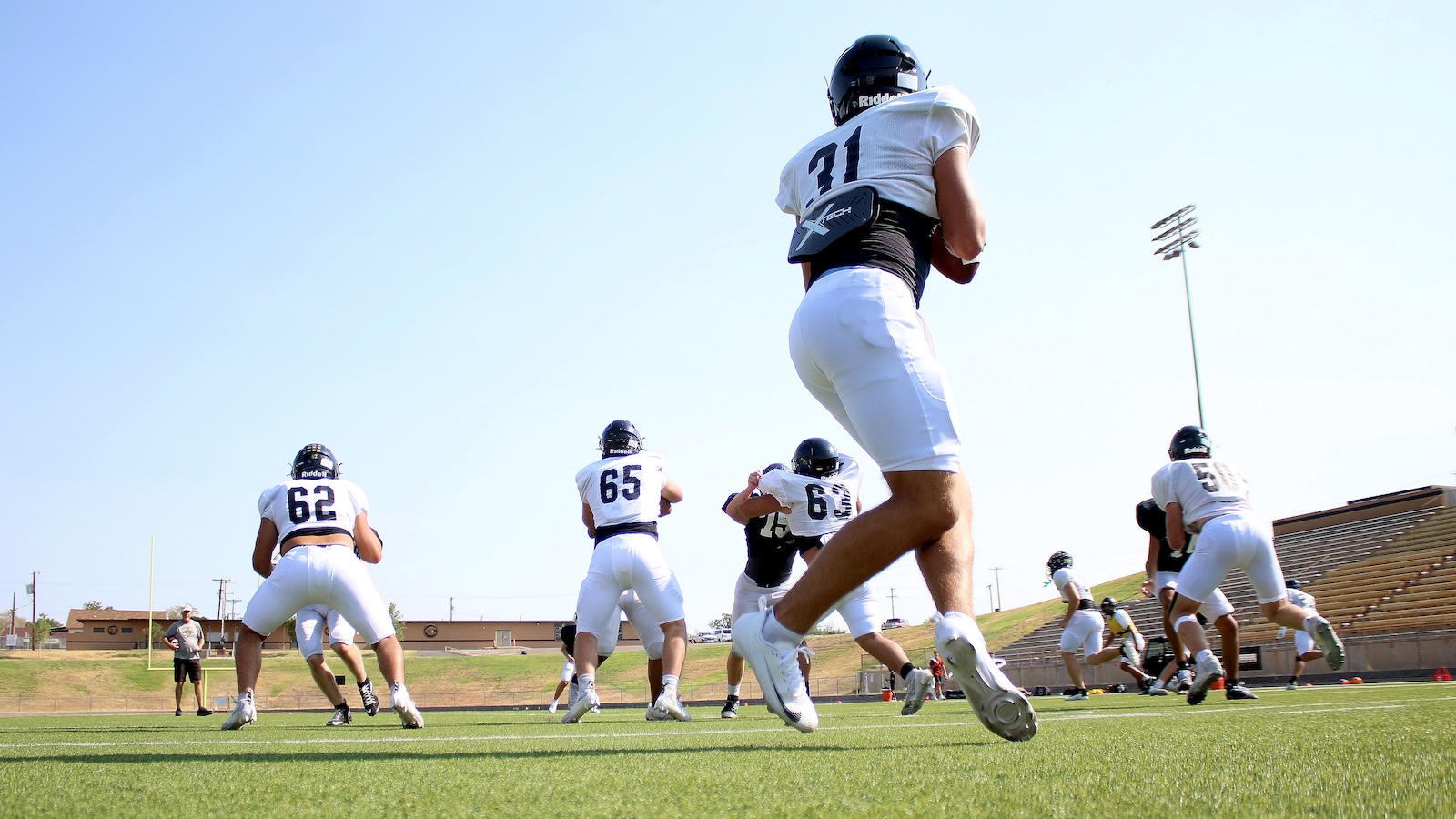Football players in white jerseys sprint on a practice field under a clear sky, number 31 in the foreground with helmet and pads on.