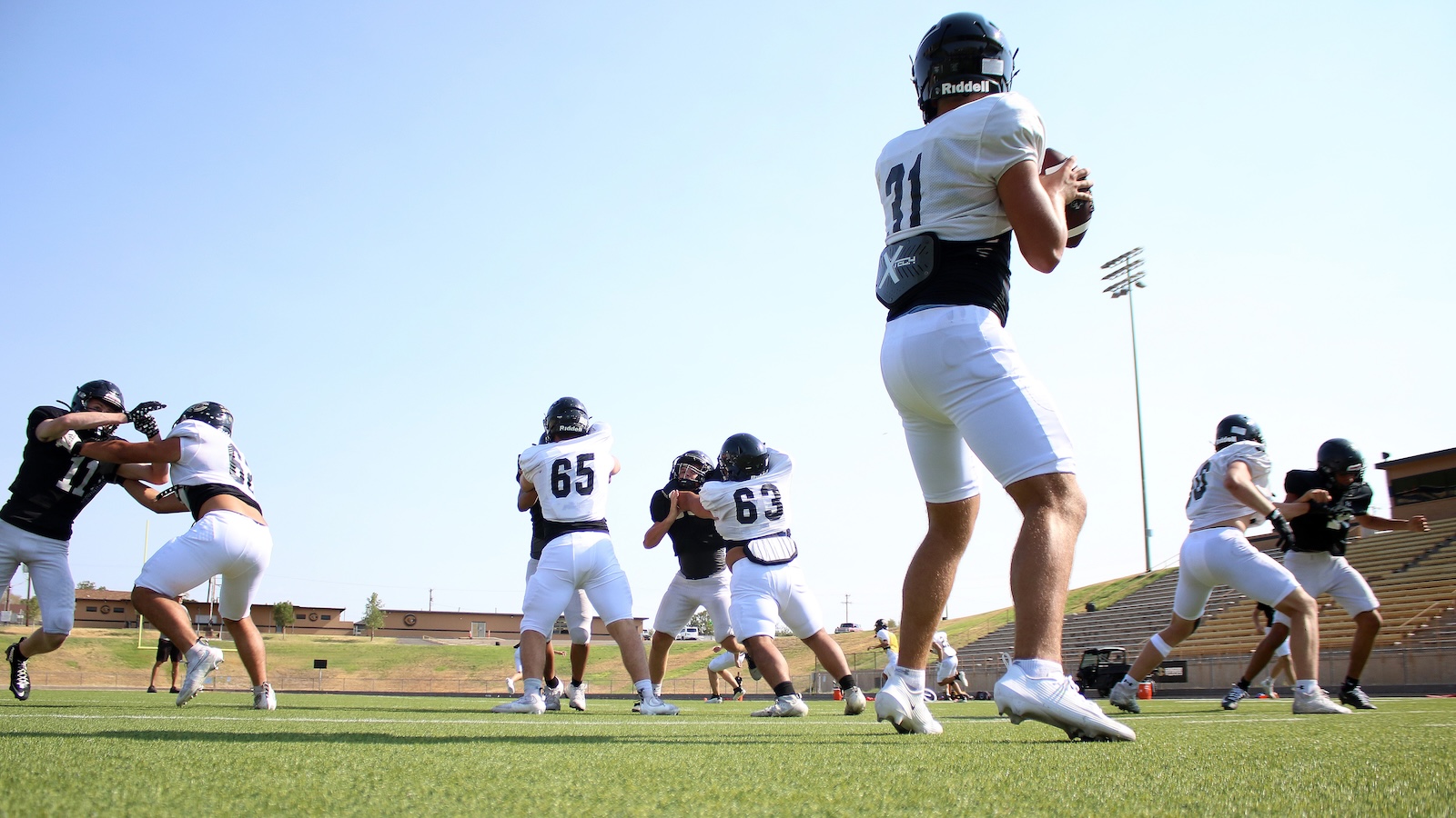 Football practice on a sunny field with players in white jerseys tackling and dodging in the foreground and background