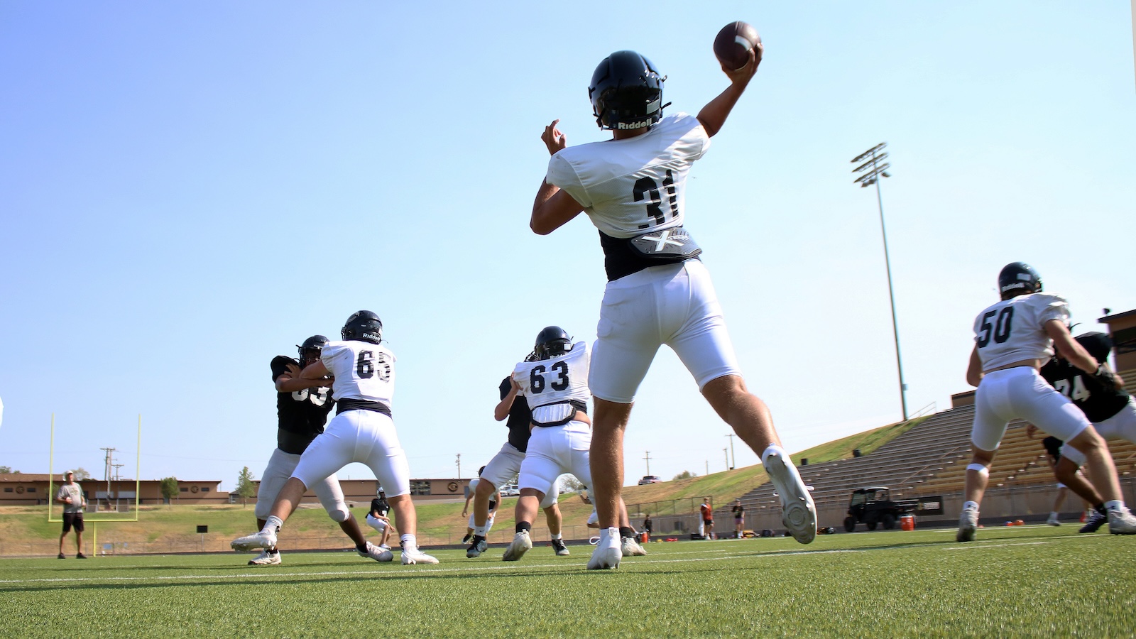 Football players in white uniforms on a sunny practice field; a player leaps with arm raised to catch a pass while teammates block nearby.