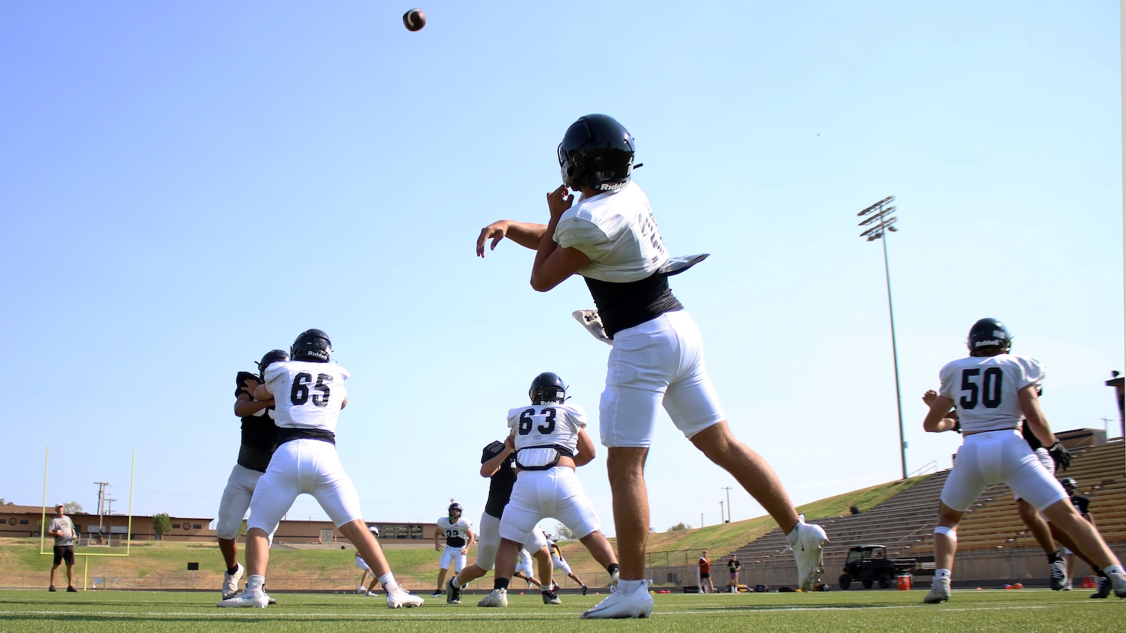 Football practice: quarterback in mid-throw with teammates blocking on a sunny field.