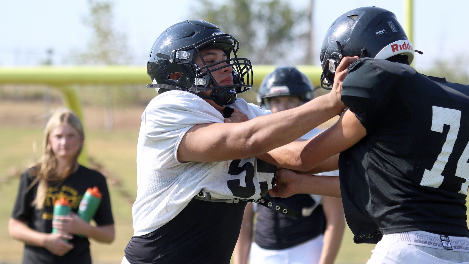 Two football players grapple during a drill, white jersey 53 against black jersey 74 on a sunny practice field.