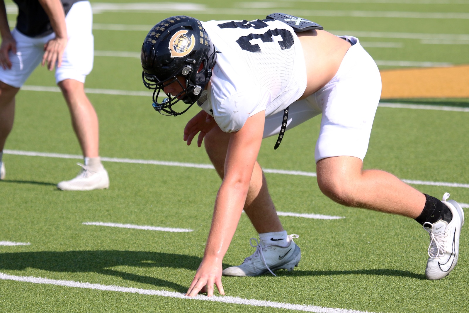 Football lineman in white uniform crouches on the turf, ready to snap the ball, helmet on with team logo.