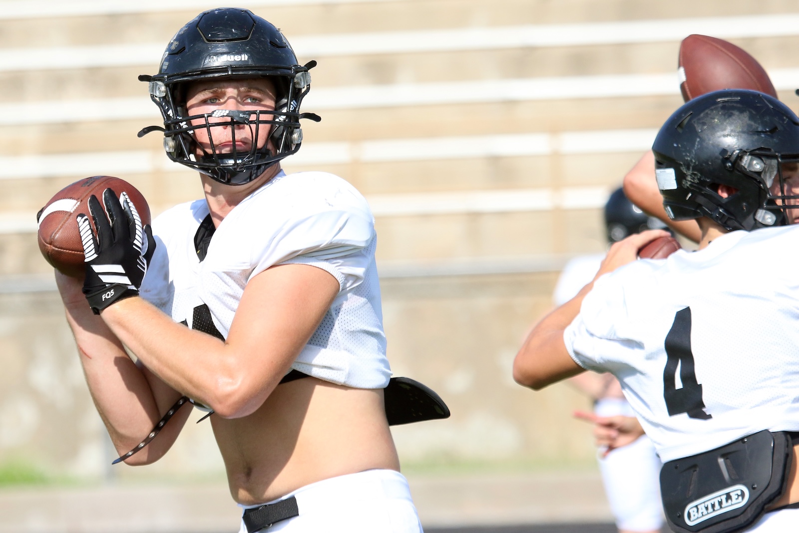 Quarterback in a white jersey grips a football, preparing to pass during a practice session with teammates in the background.