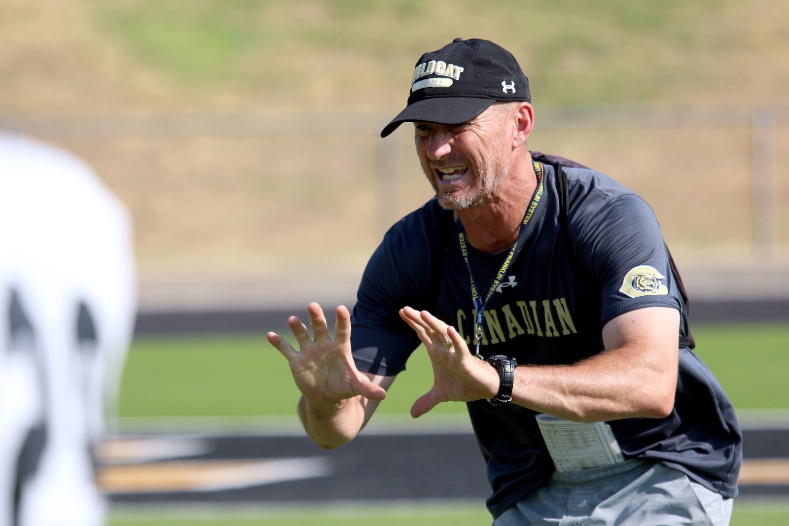 Athletic coach in a black cap and dark shirt shouting and guiding a drill on a track field.