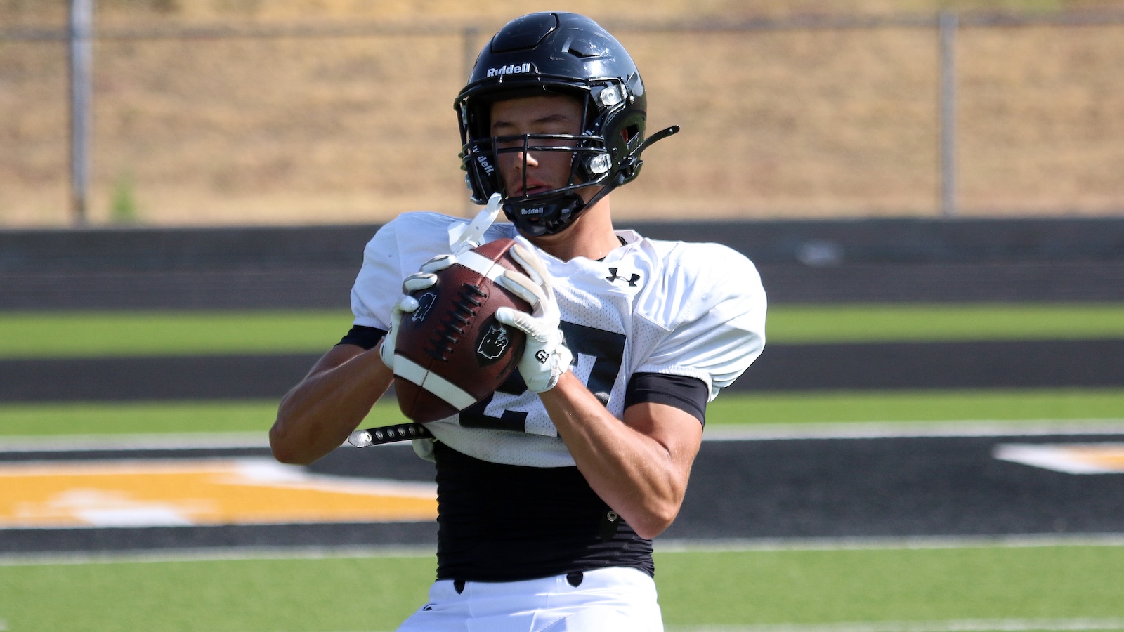 Football player in a white jersey and black helmet holding a football close to his chest on the field at practice/game moment.