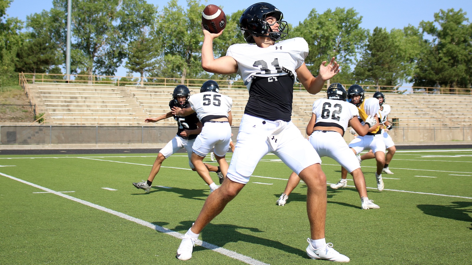 Football quarterback in white and black uniform throws a pass on a sunny outdoor field, teammates and defenders in the background.