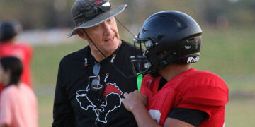 Two people on a grassy field share a hug; one in a black shirt with a graphic, gray hat and sunglasses, the other in a red jersey with a black helmet.