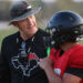 Two people on a grassy field share a hug; one in a black shirt with a graphic, gray hat and sunglasses, the other in a red jersey with a black helmet.