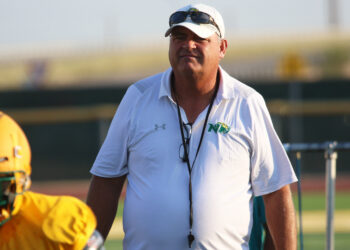Coach wearing a white polo and cap with sunglasses stands on a football field, yellow helmet visible to the left.