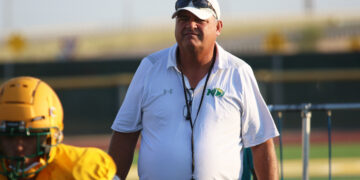 Coach wearing a white polo and cap with sunglasses stands on a football field, yellow helmet visible to the left.