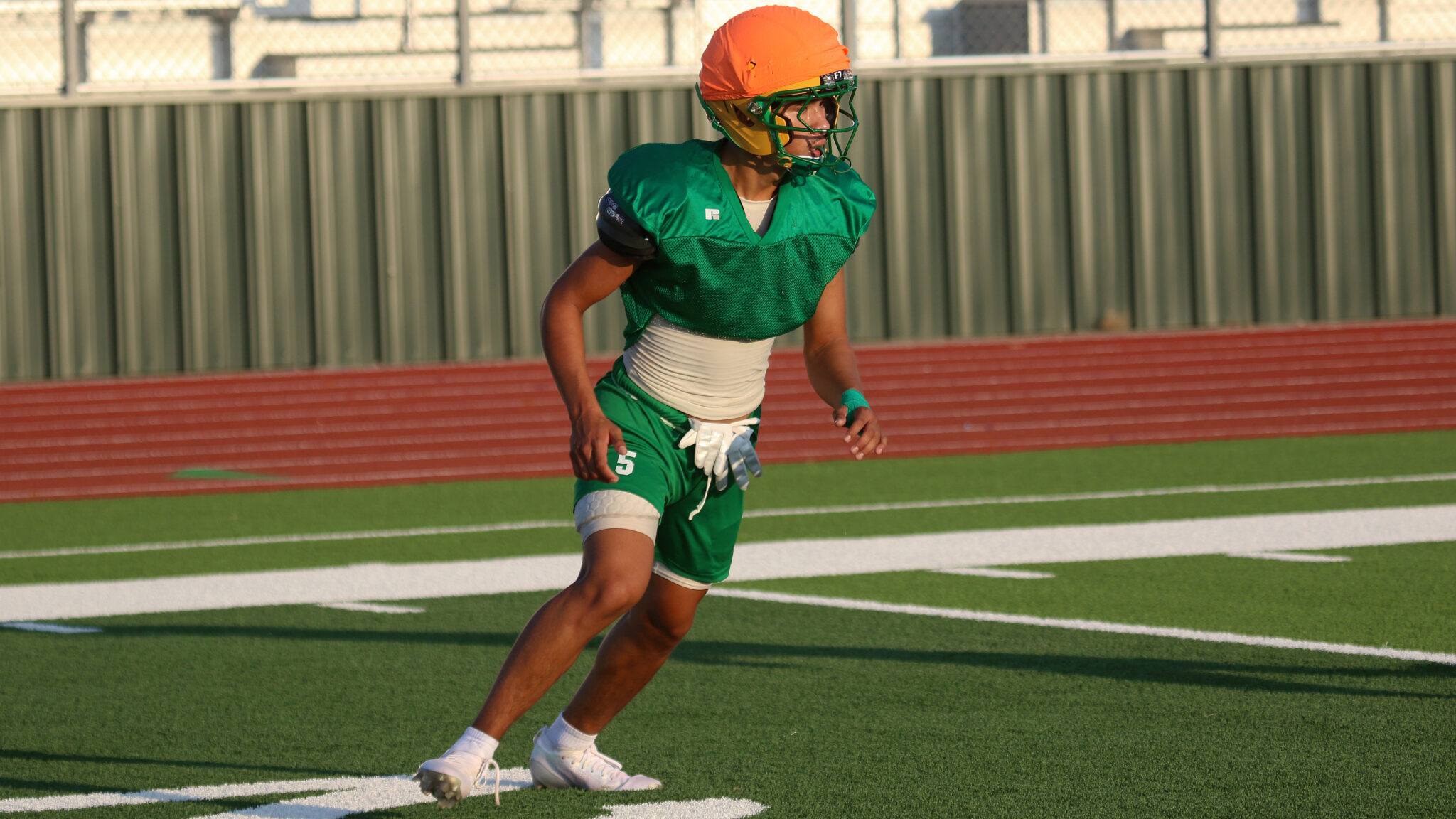 New Deal - Josiah Mendoza 2 | Press Pass Sports Football player in green jersey and orange helmet runs on a track- turf field with a fence in the background.