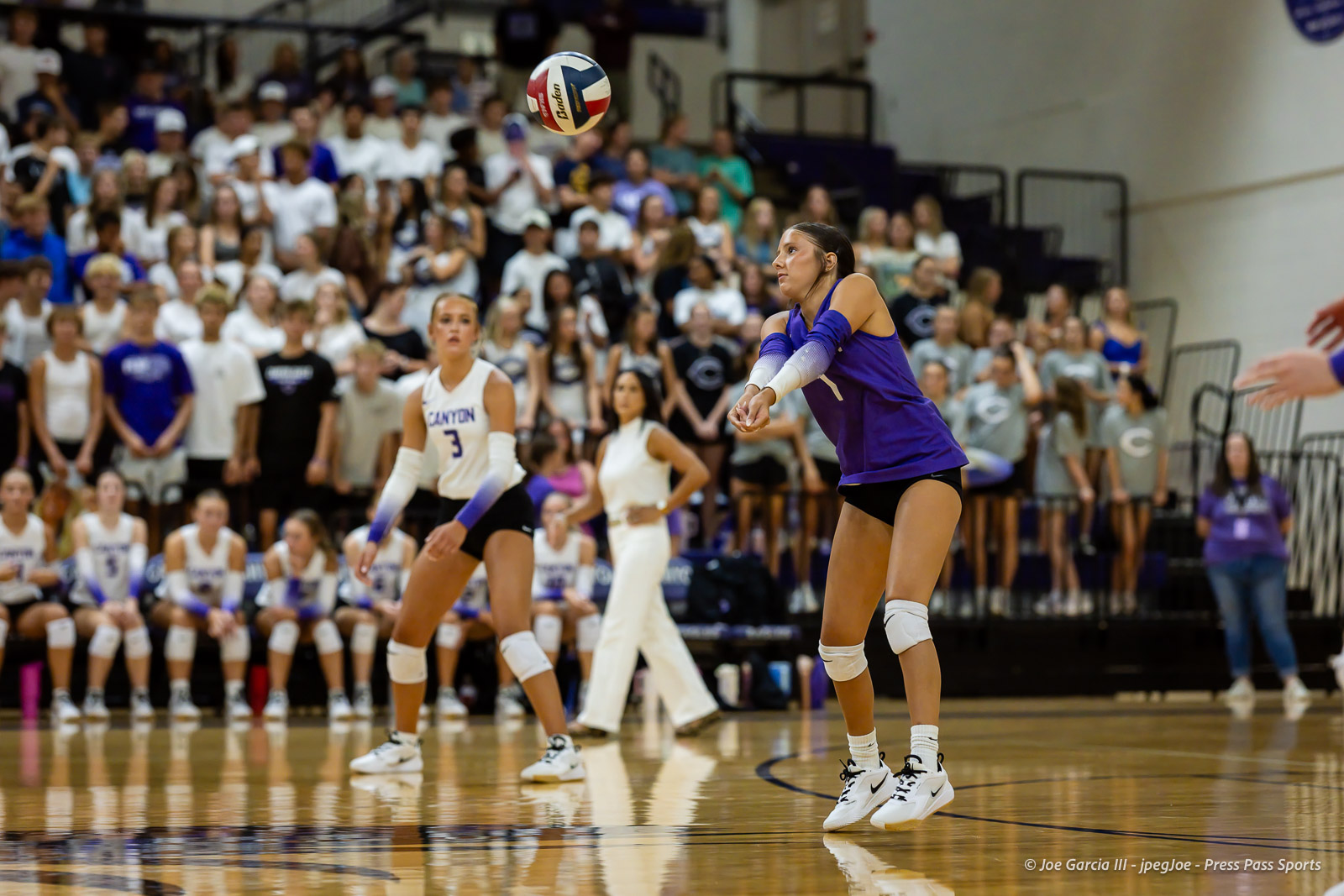 Female volleyball players in purple and white uniforms on a gym court, ball in midair during a rally with a crowded crowd in the stands behind them.