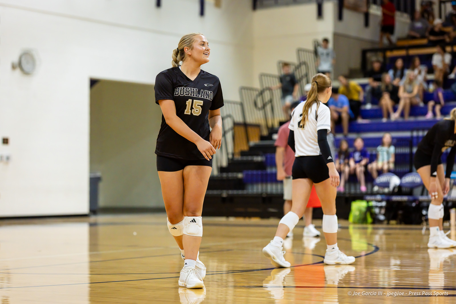 Smiling female volleyball player in a black jersey number 15 walks on a gym floor during a match, with teammates and spectators in the bleachers behind her.
