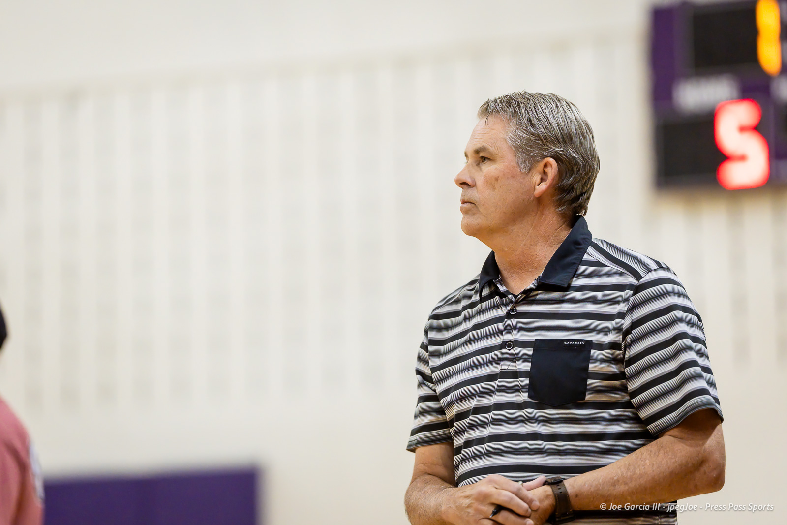 Side profile of a middle-aged man with gray hair wearing a striped polo, standing in a gym indoor setting near a scoreboard in the background.