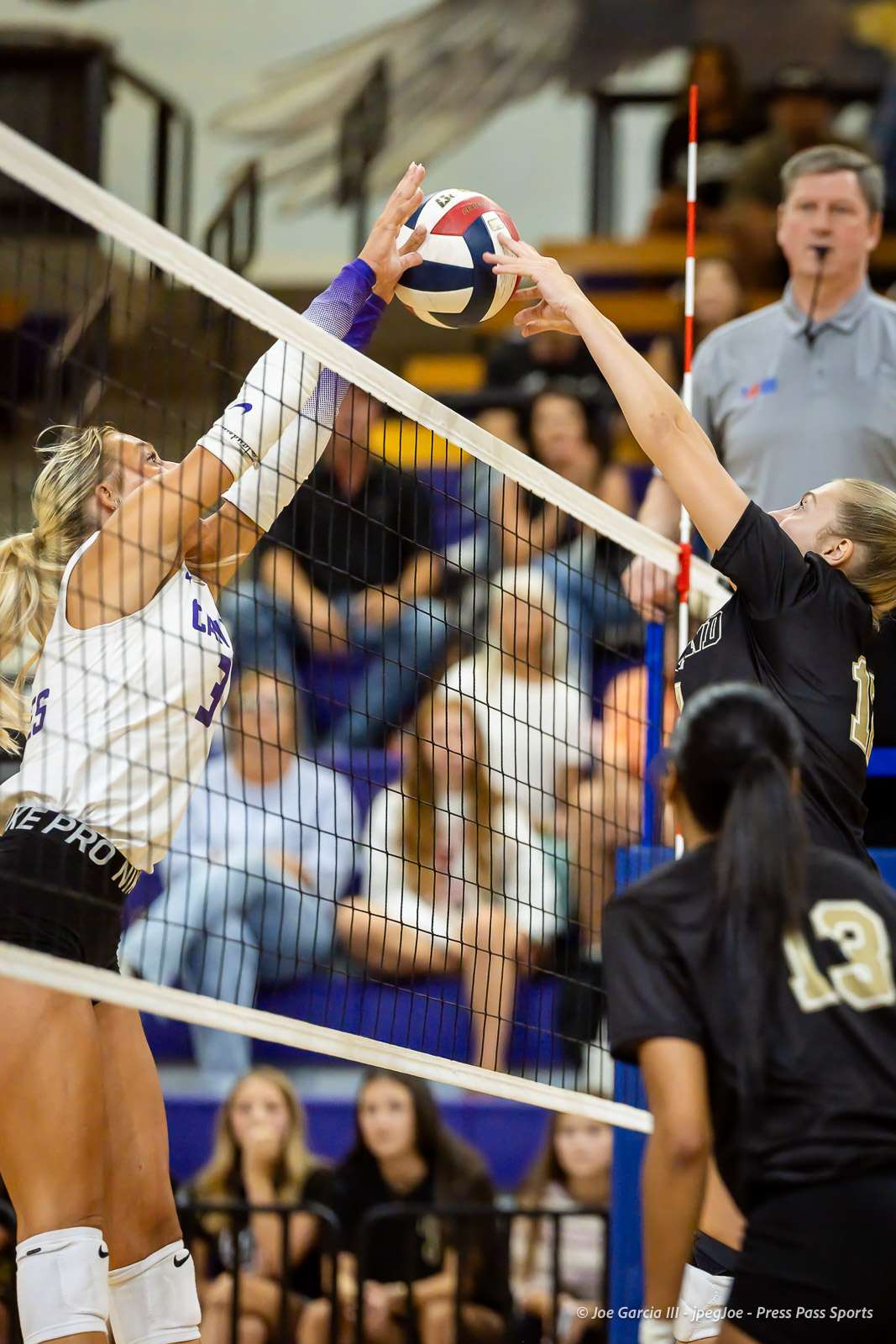 Two female players leap at the net, hands colliding to block a volleyball as it hovers between them.