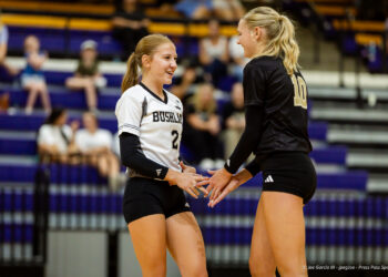 Two volleyball players share a high-five on the court after a point; white jersey number 2 and black jersey number 10, with fans in bleachers behind them.