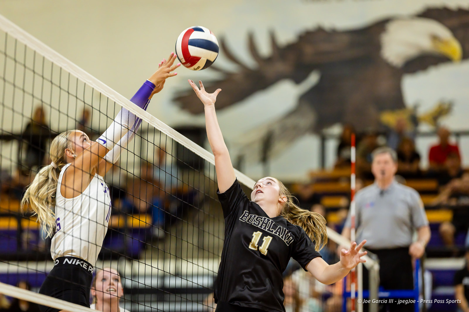 Two female volleyball players jump at the net reaching for a ball during a rally.