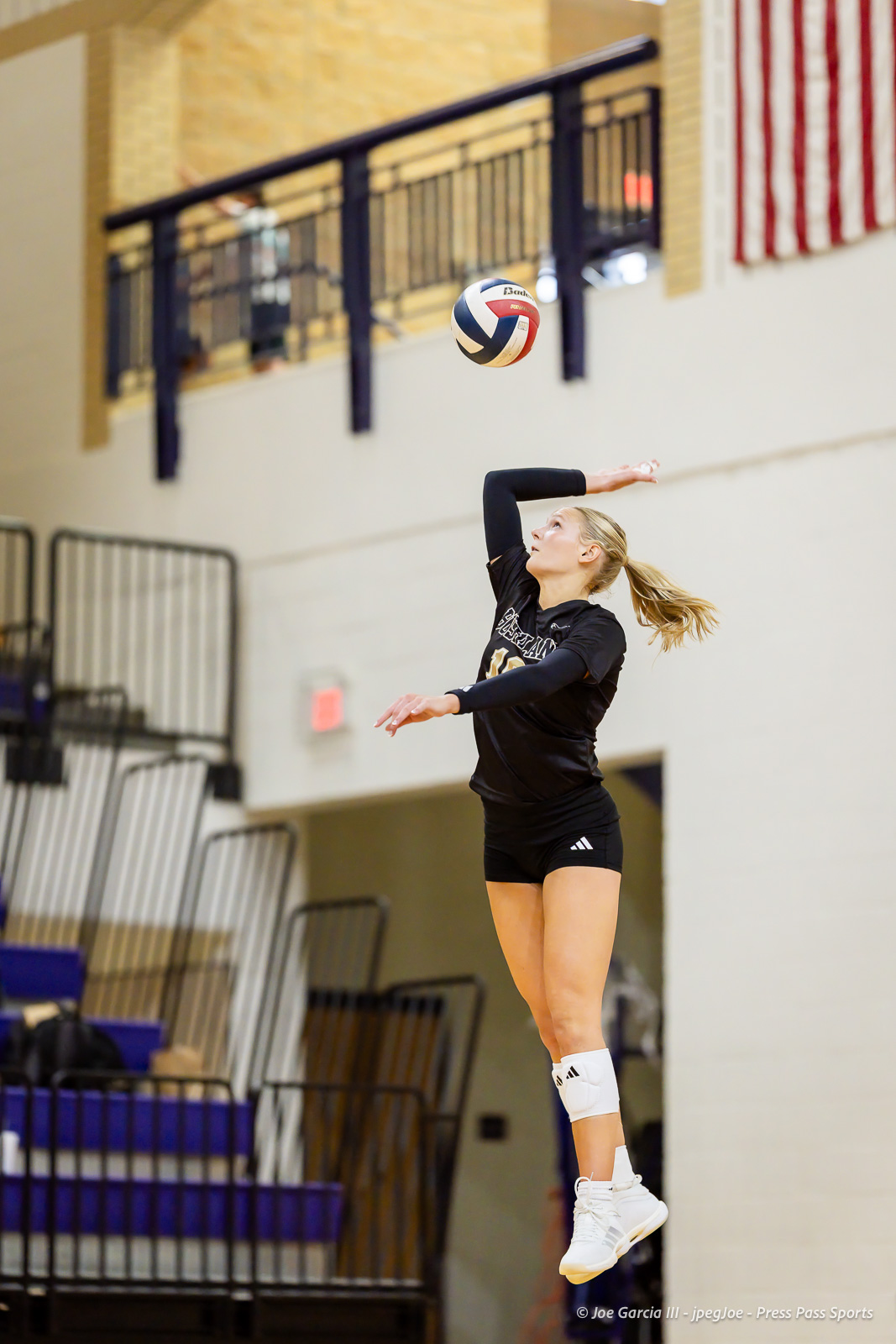 Female volleyball player in mid-air jump, serving or spiking the ball in a gym with an American flag in the background.