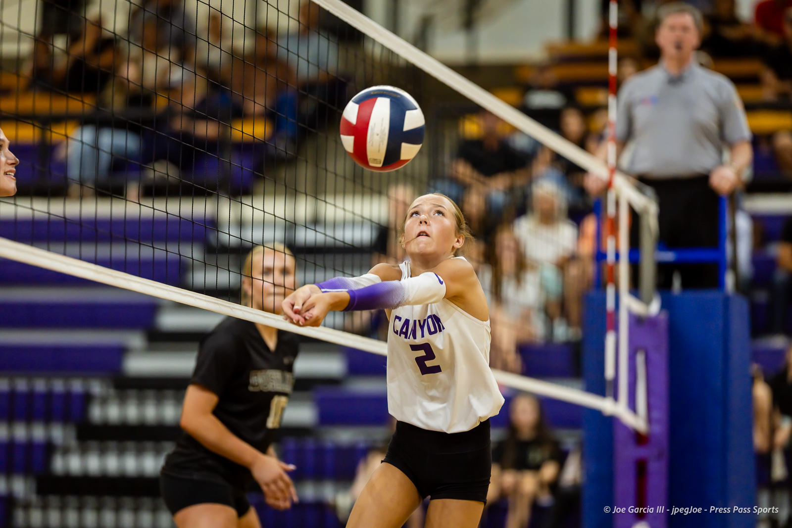 Female volleyball player in white jersey #2 lunges toward the ball at the net during a match; crowd seated in the stands behind her.