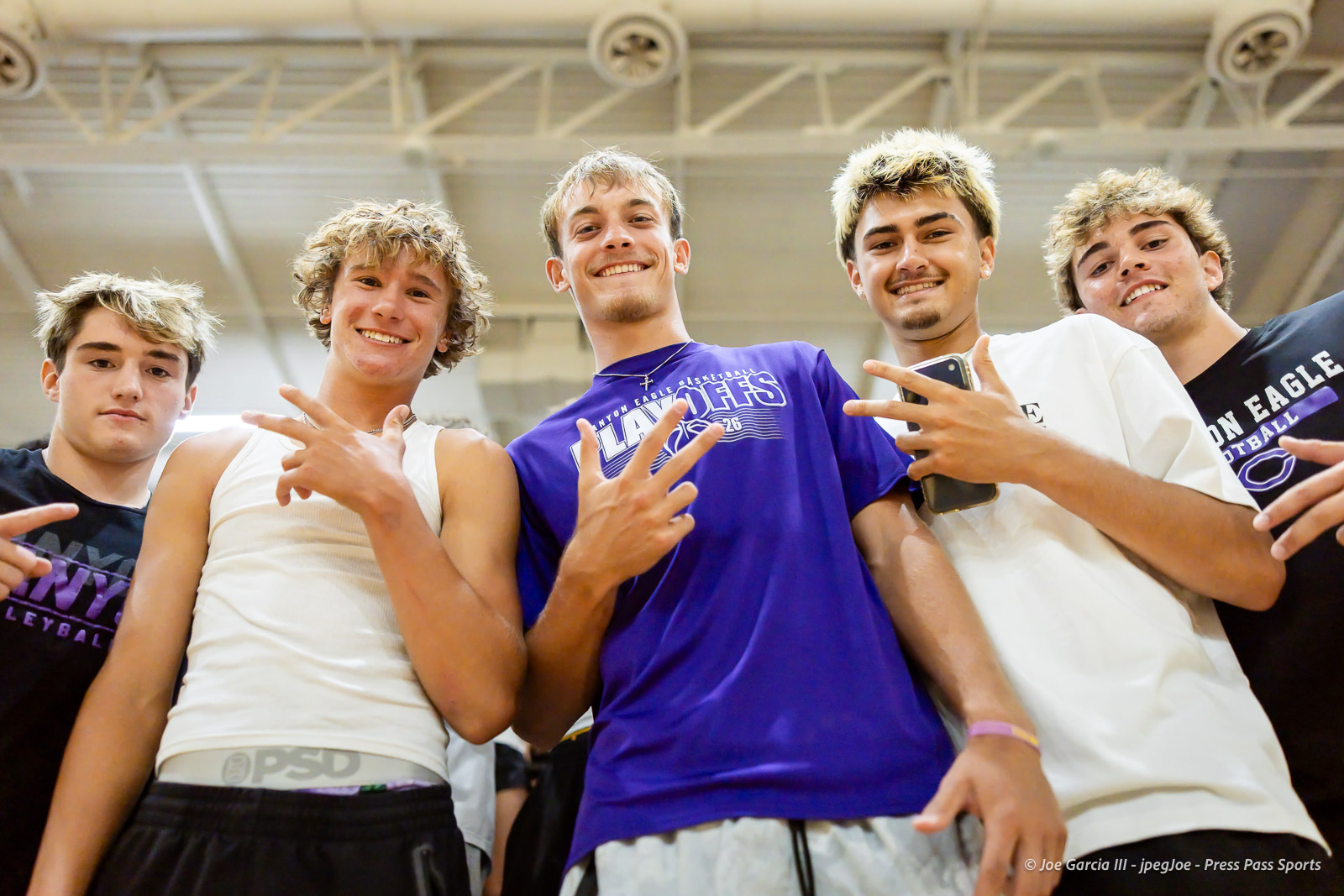 Five young men posing together in a gym, smiling and making hand signs, wearing basketball team shirts.