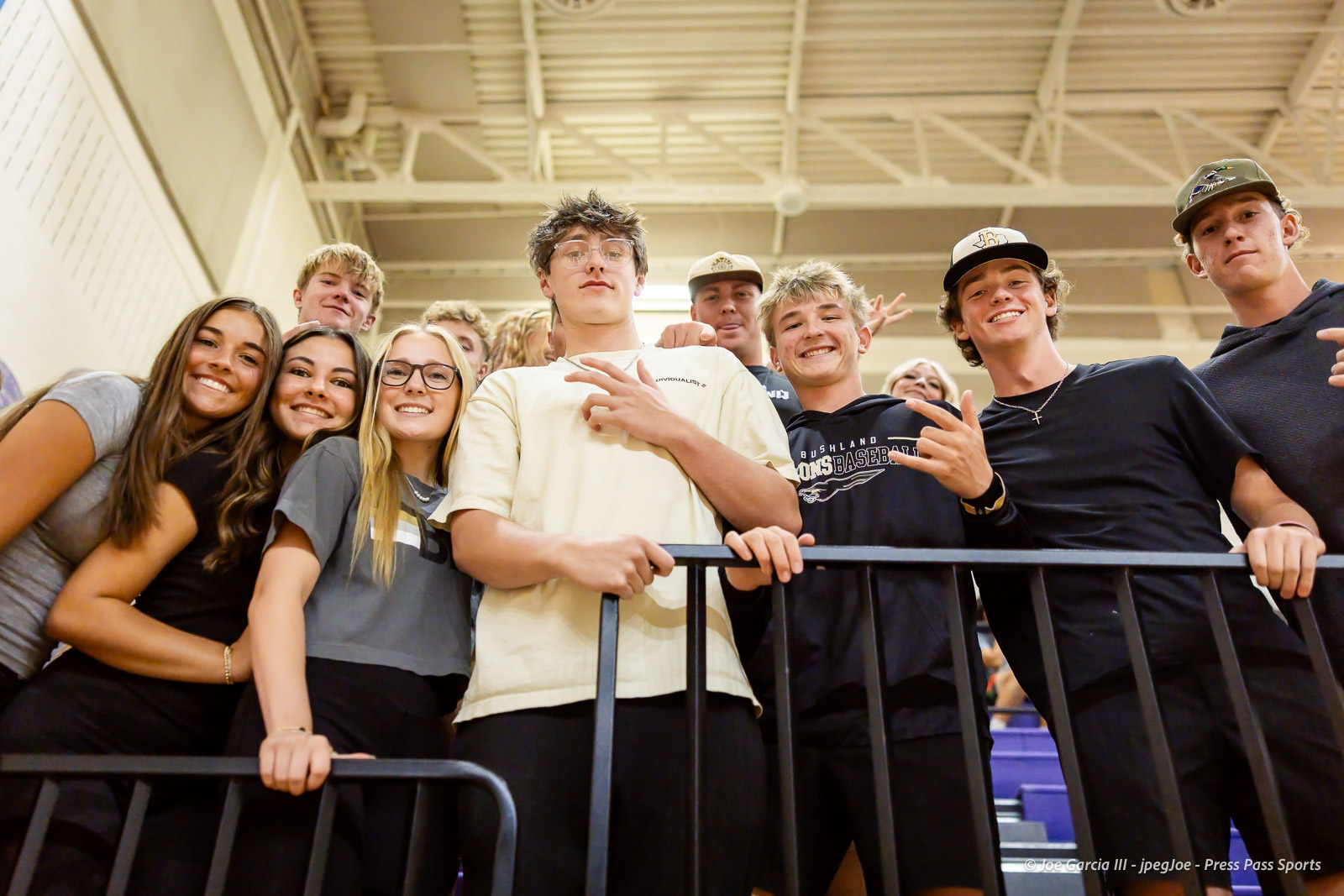Group of smiling teenagers posing at a railing in a gym or stadium, looking toward the camera.