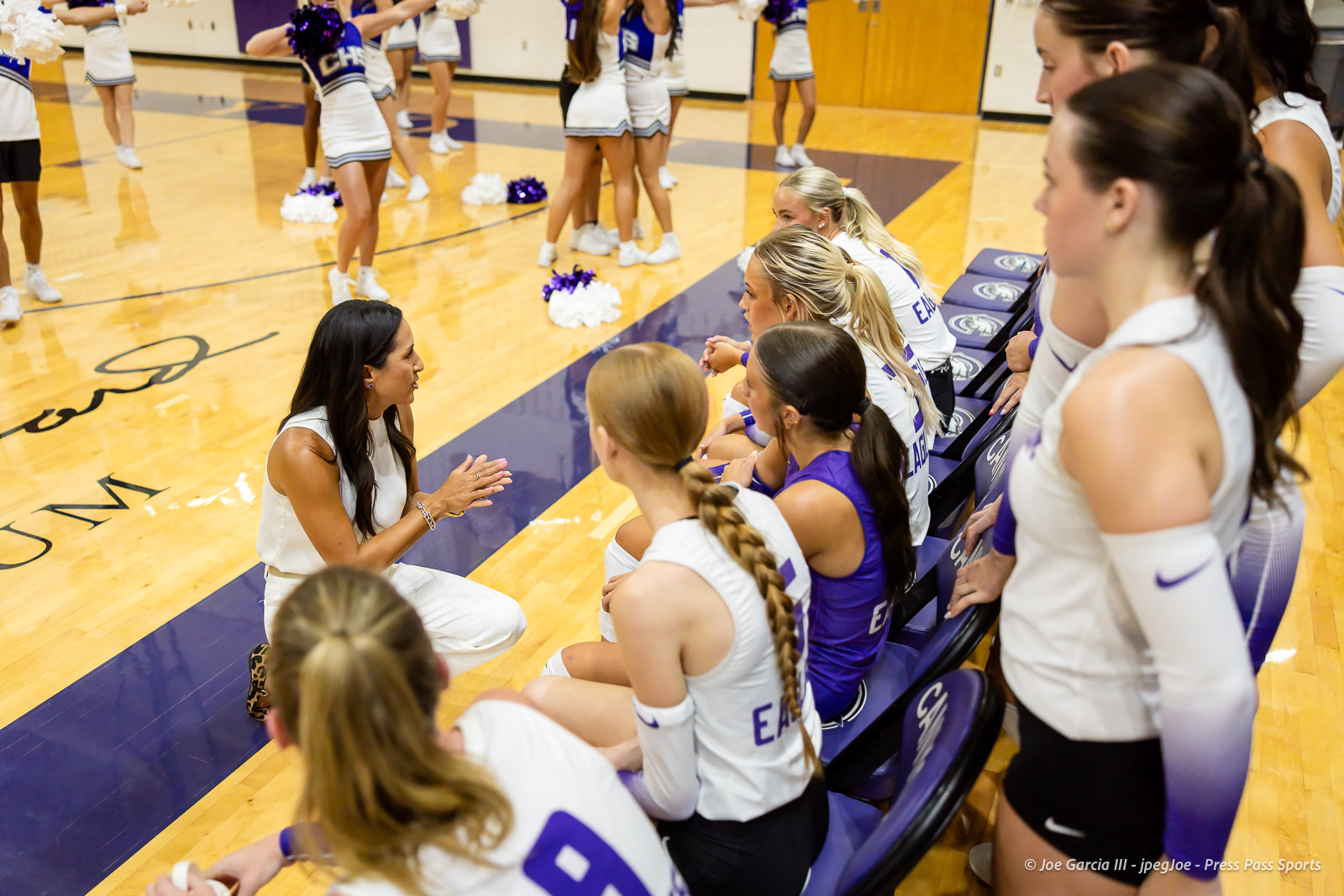 Female coach kneels to address seated basketball players in white and purple uniforms on the bench during a game in the gym.