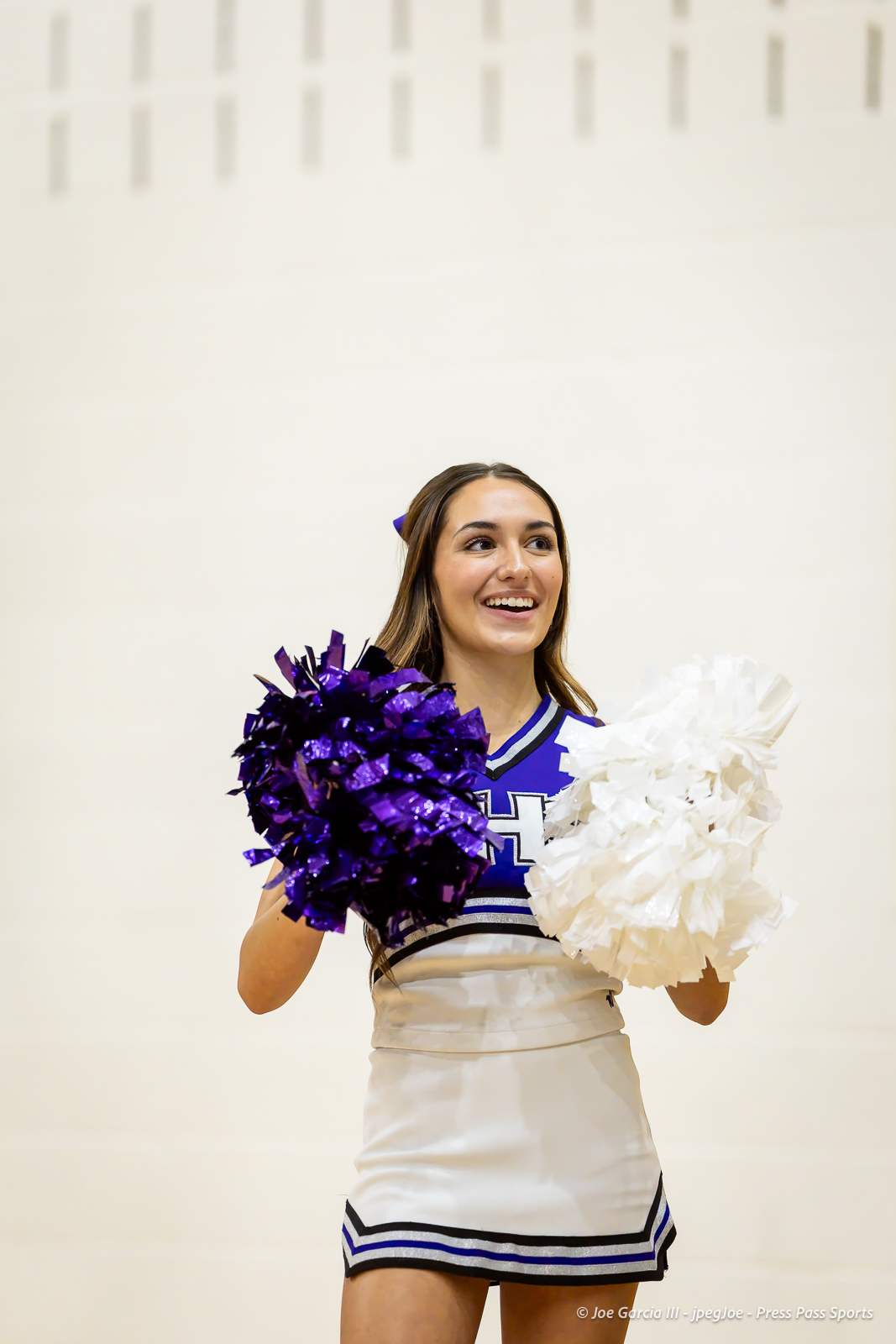 Cheerleader in blue and white uniform smiles, holding purple pom-poms in each hand.