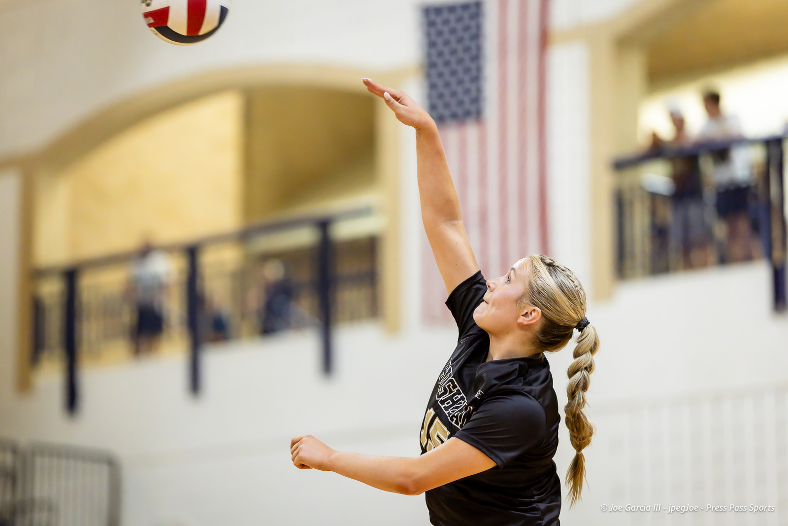 Female volleyball player in a black shirt spikes an indoor volleyball with her arm extended upward.
