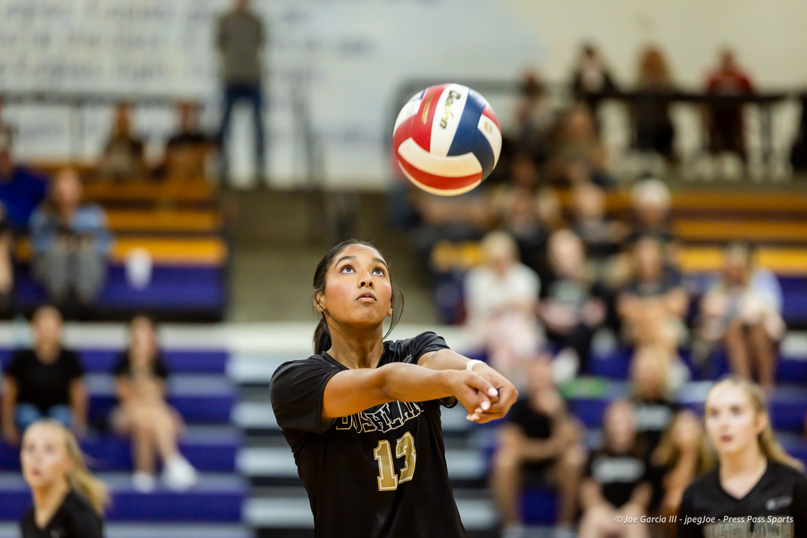 Female volleyball player in a black jersey (#13) sets the ball with forearms as it floats above her, with a gym full of spectators in the background.