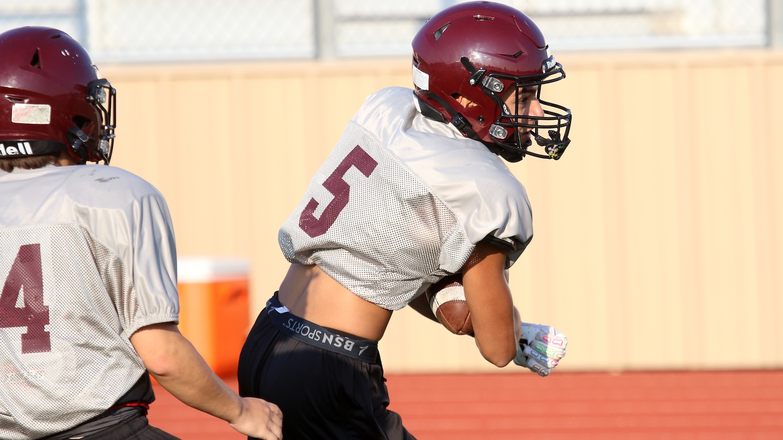Two football players in maroon helmets and gray jerseys sprinting on a track; one carries a ball tucked under his arm while a teammate runs beside him.