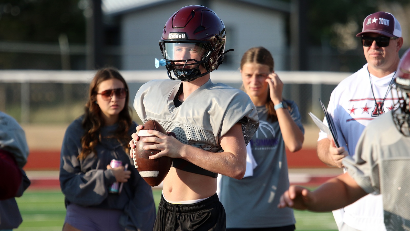 Youth football player in gray jersey and maroon helmet holding a football on a practice field, teammates and a coach in the background.