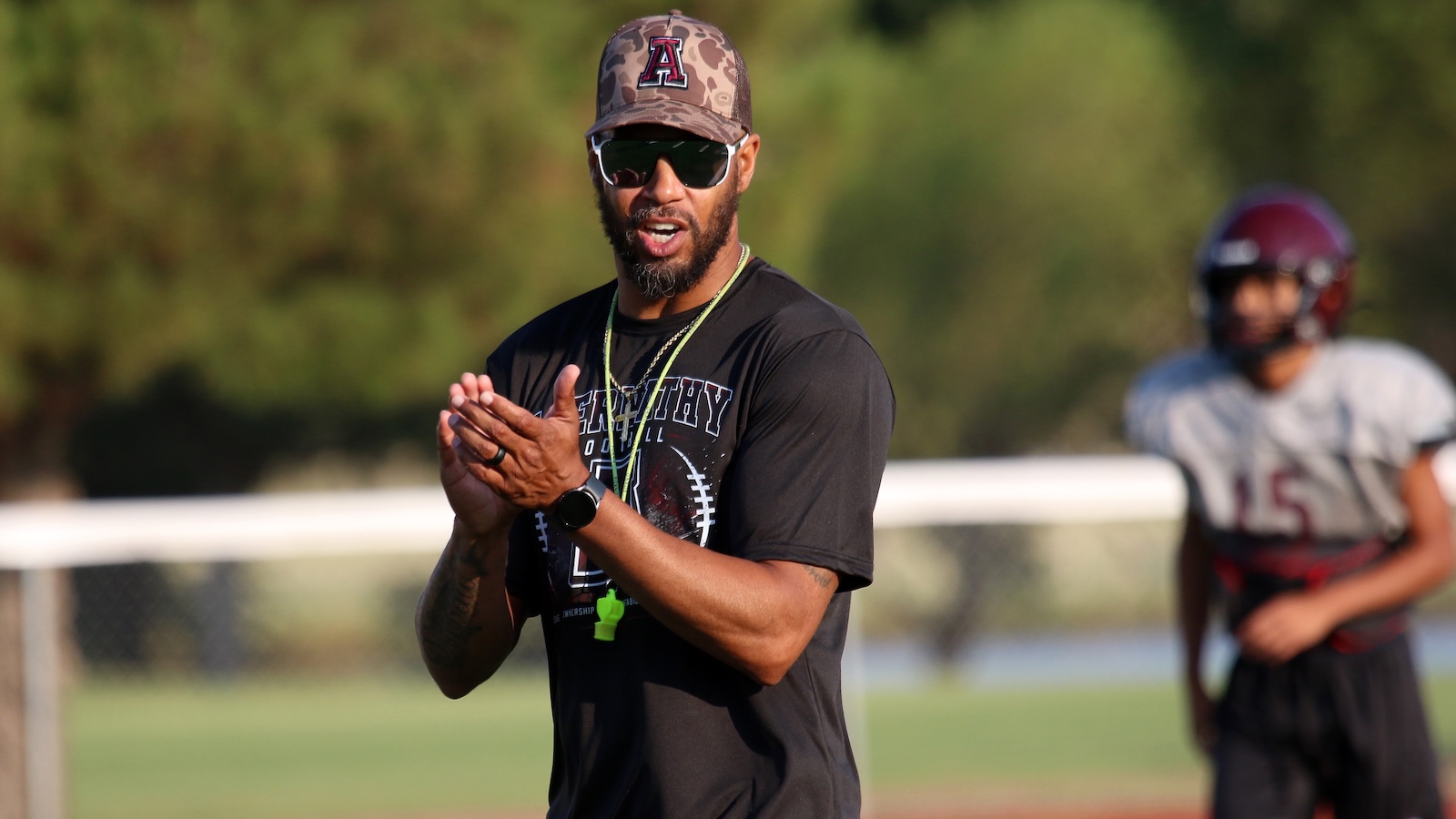 Coach clapping on a sunny football field, with a player in a helmet in the background.