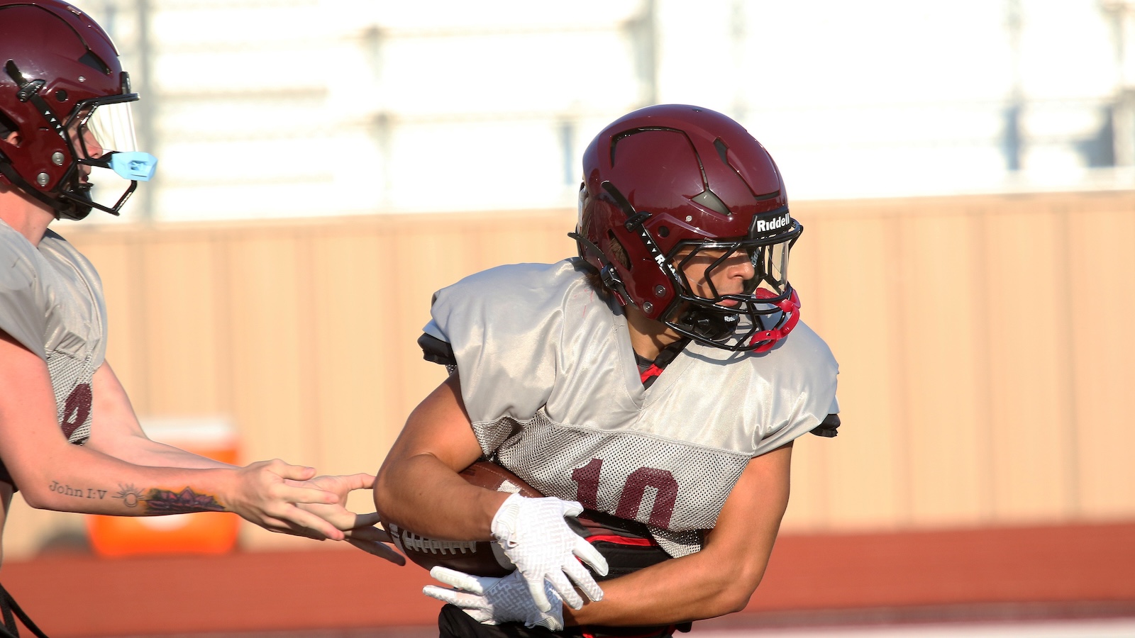 Two football players in maroon helmets and gray practice jerseys compete for the ball on a field; one wears number 10 on his jersey.