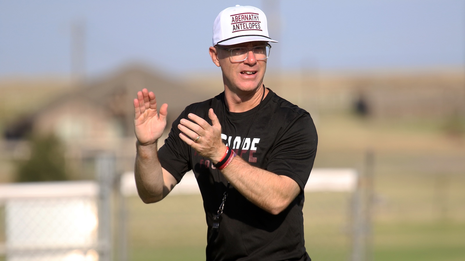 Man in a black t-shirt and glasses applauding outdoors, wearing a white cap that says'ABERNATHY ANTELOPES' at a sunny field.