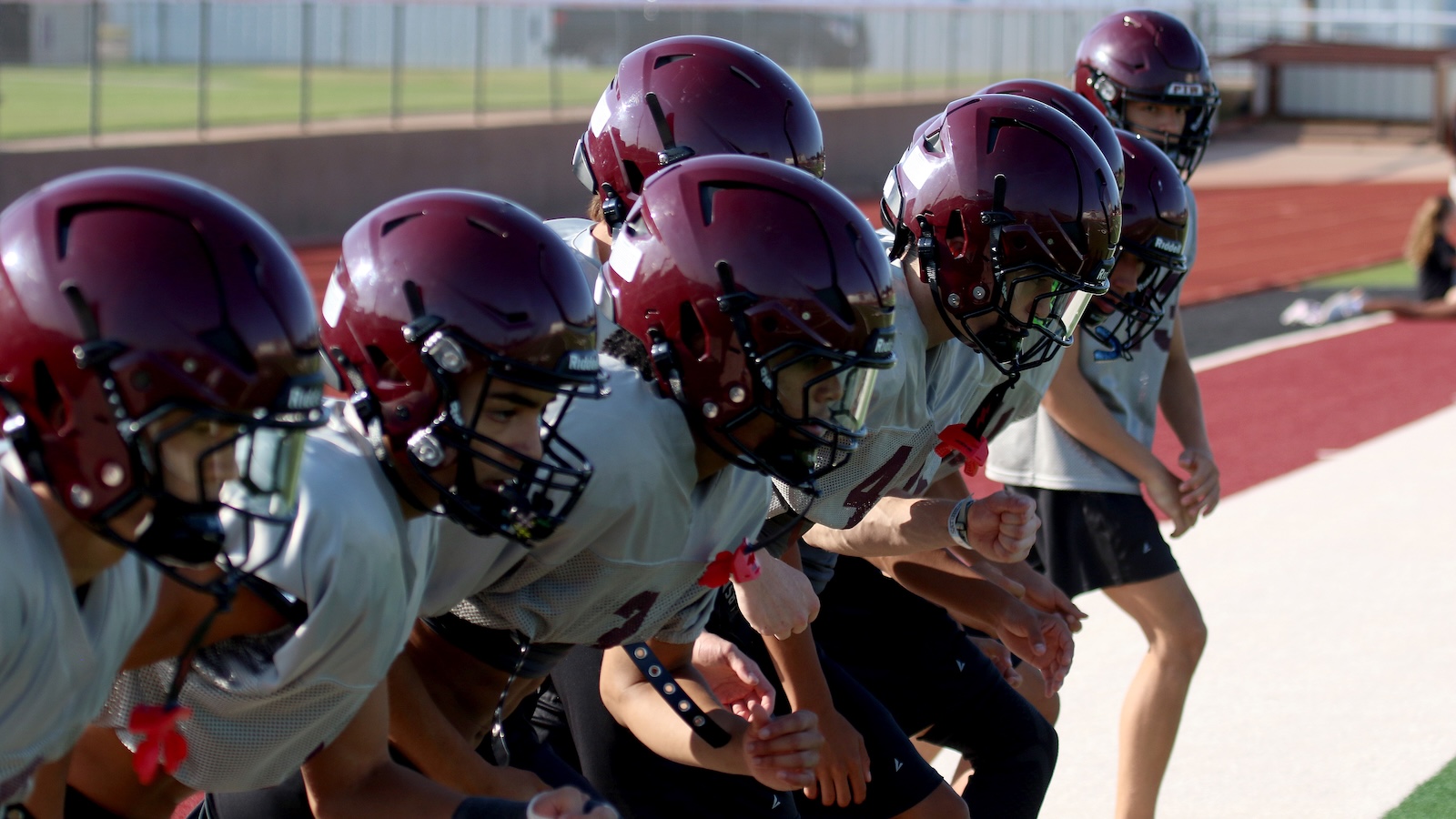 Football players in maroon helmets crouch at the line of scrimmage on a track field, ready to start the play.