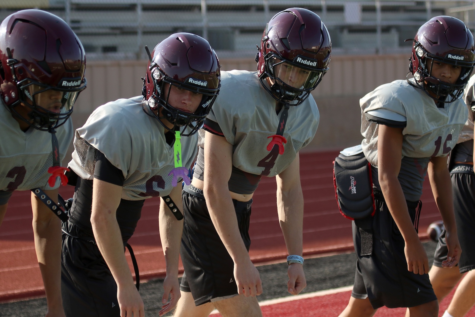 Four football players wearing maroon helmets and gray jerseys line up at the start on a track field, ready to snap.