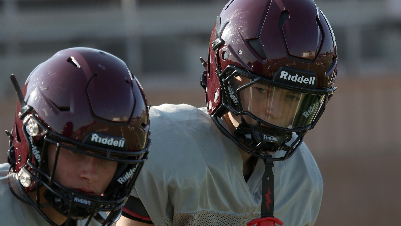 Two football players in maroon Riddell helmets during practice, one facing the camera with a focused expression.