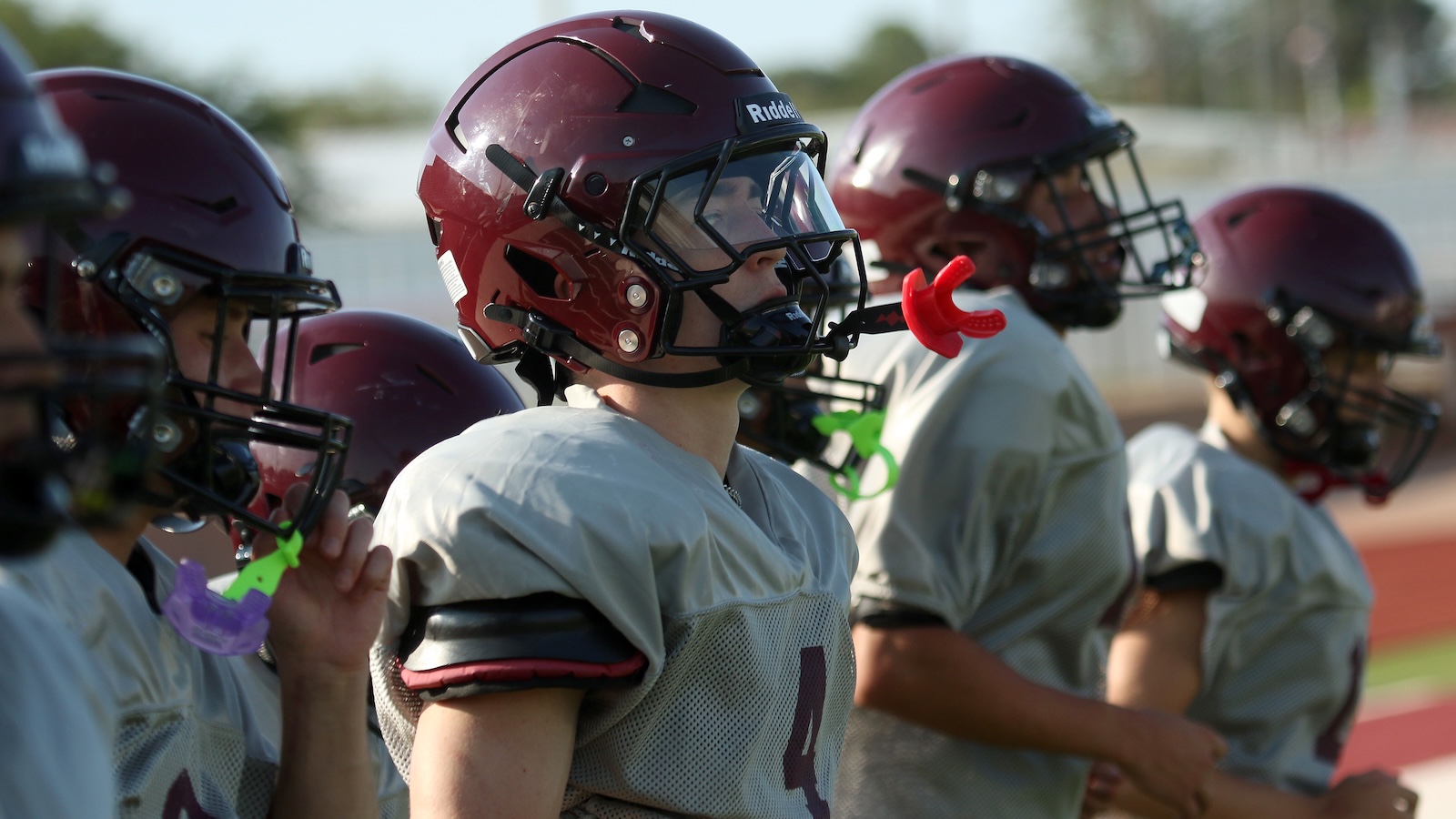 Youth football players in maroon helmets and gray practice jerseys stand on the sideline during a game or drill.