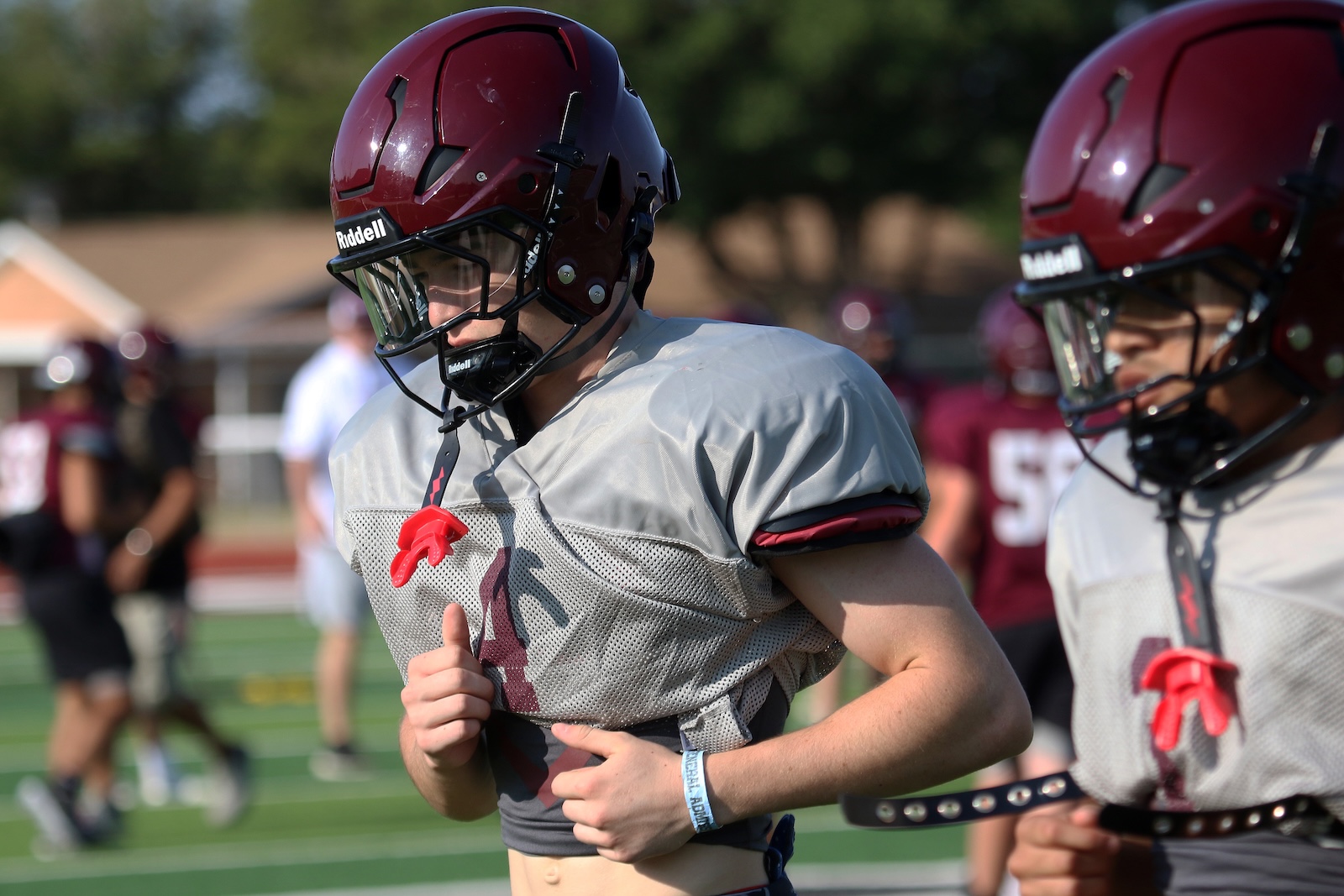 Two football players in maroon helmets and gray practice jerseys jog on a sunny field, foreground player adjusting his jersey strap.