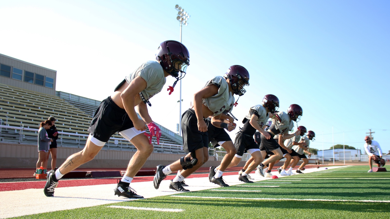 Group of football players in helmets and pads line up at the starting line on a track, ready to sprint during practice on a sunny day.