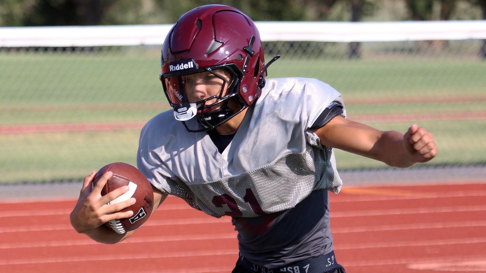 Young football player in a gray practice jersey and maroon helmet runs with a football tucked under his arm on a red track field.