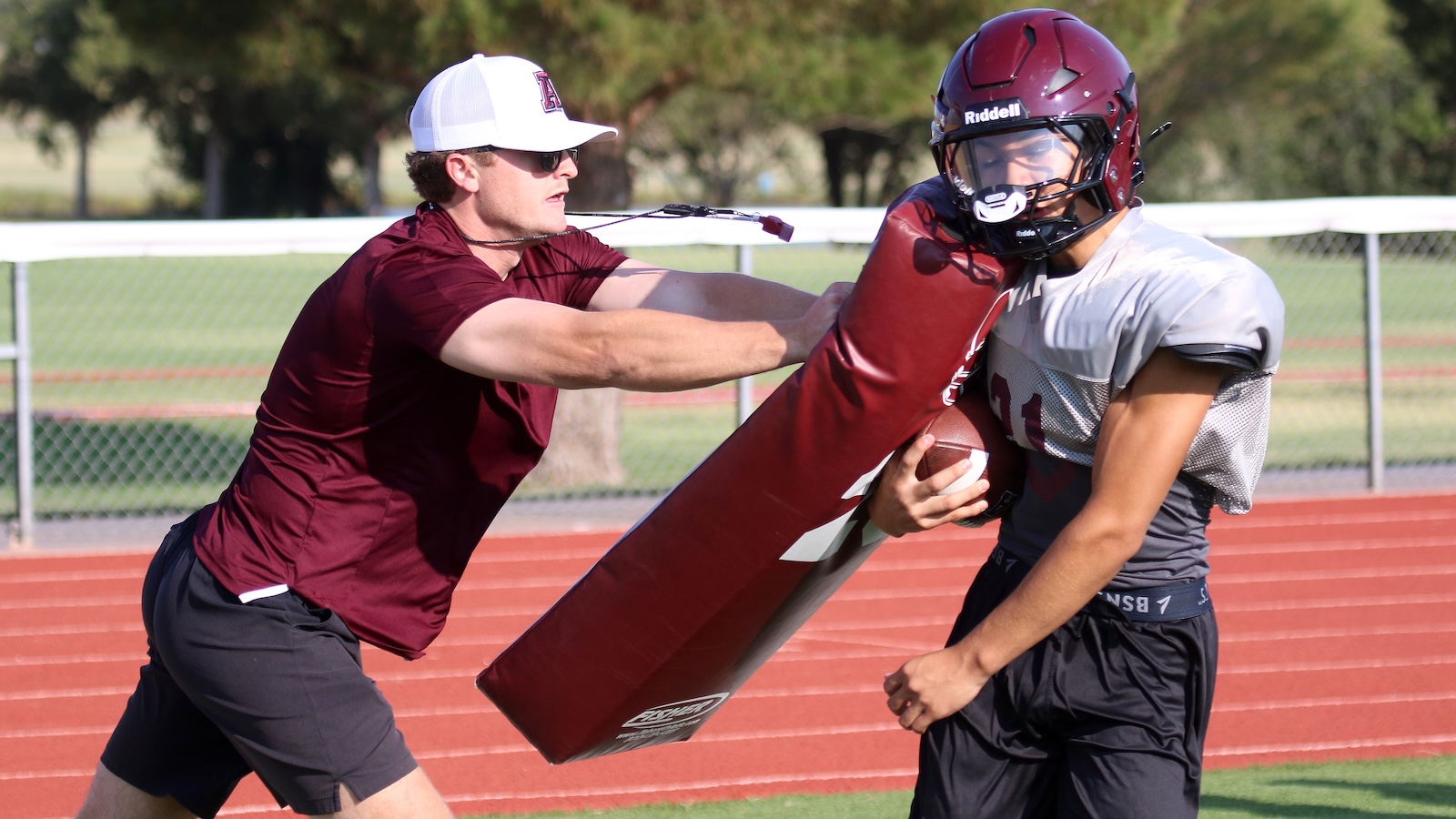 Coach in maroon shirt and white cap blocks a football player with a padded tackling dummy during practice on a track field edge edge.