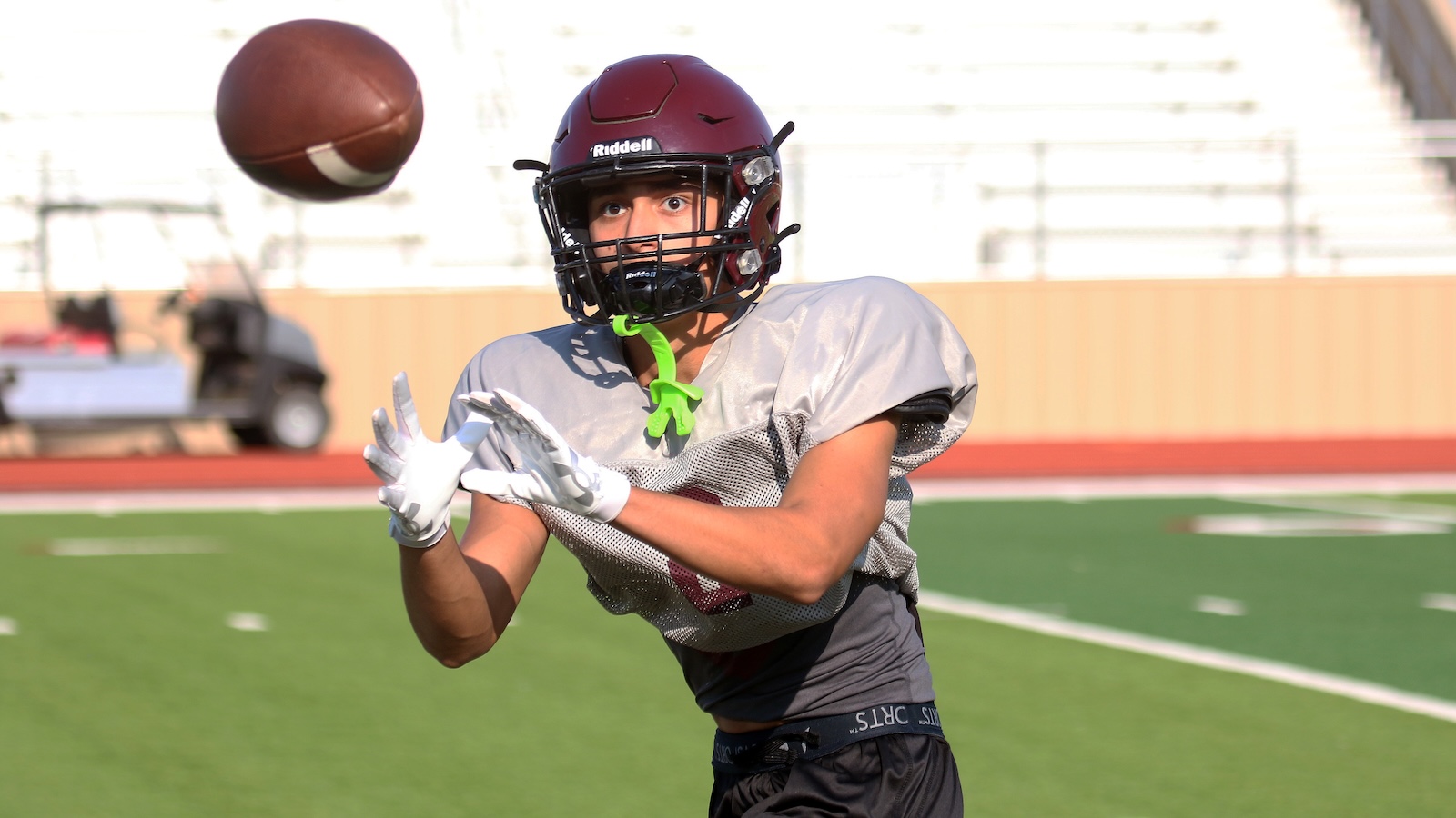 Football player in a maroon helmet and gray jersey reaches to catch a football on a green field during practice Mossy daylight